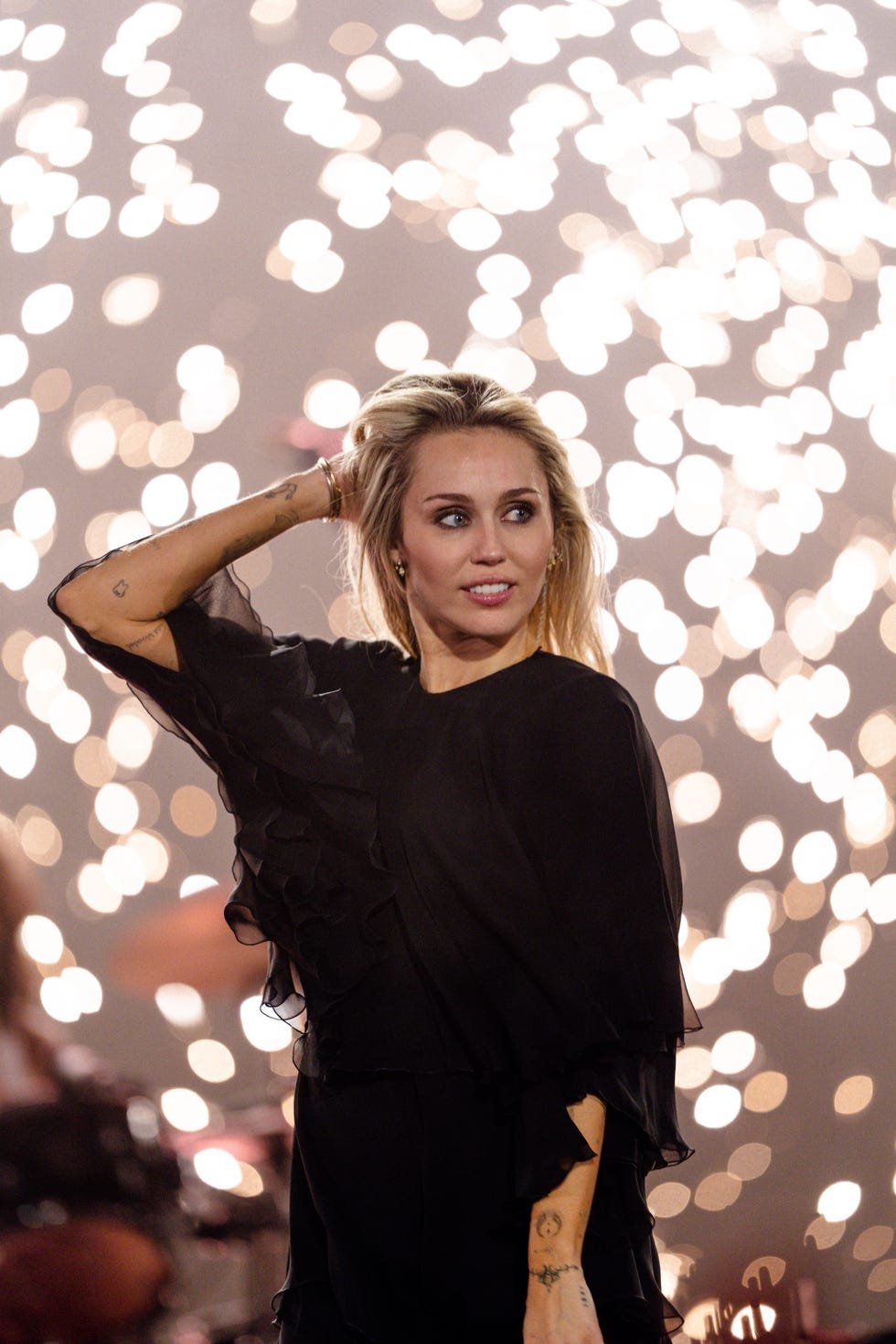 A woman in a black dress poses confidently against a backdrop of bright, blurred lights.